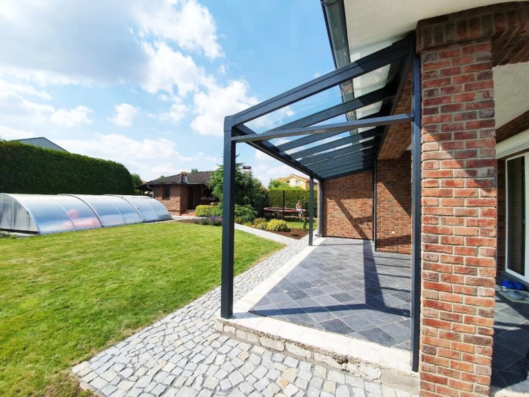 Moderná antracitová hliníková pergola so sklenenou strechou pri tehlovom dome so záhradou a bazénom.